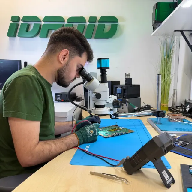 Tehnician la microscop peste o placă pe covorașul antistatic, cu penseta și stația de lipit alături